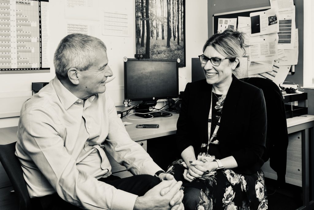A man and a woman sat on chairs chatting in an office with a desk behind them and a busy noticeboard on the wall